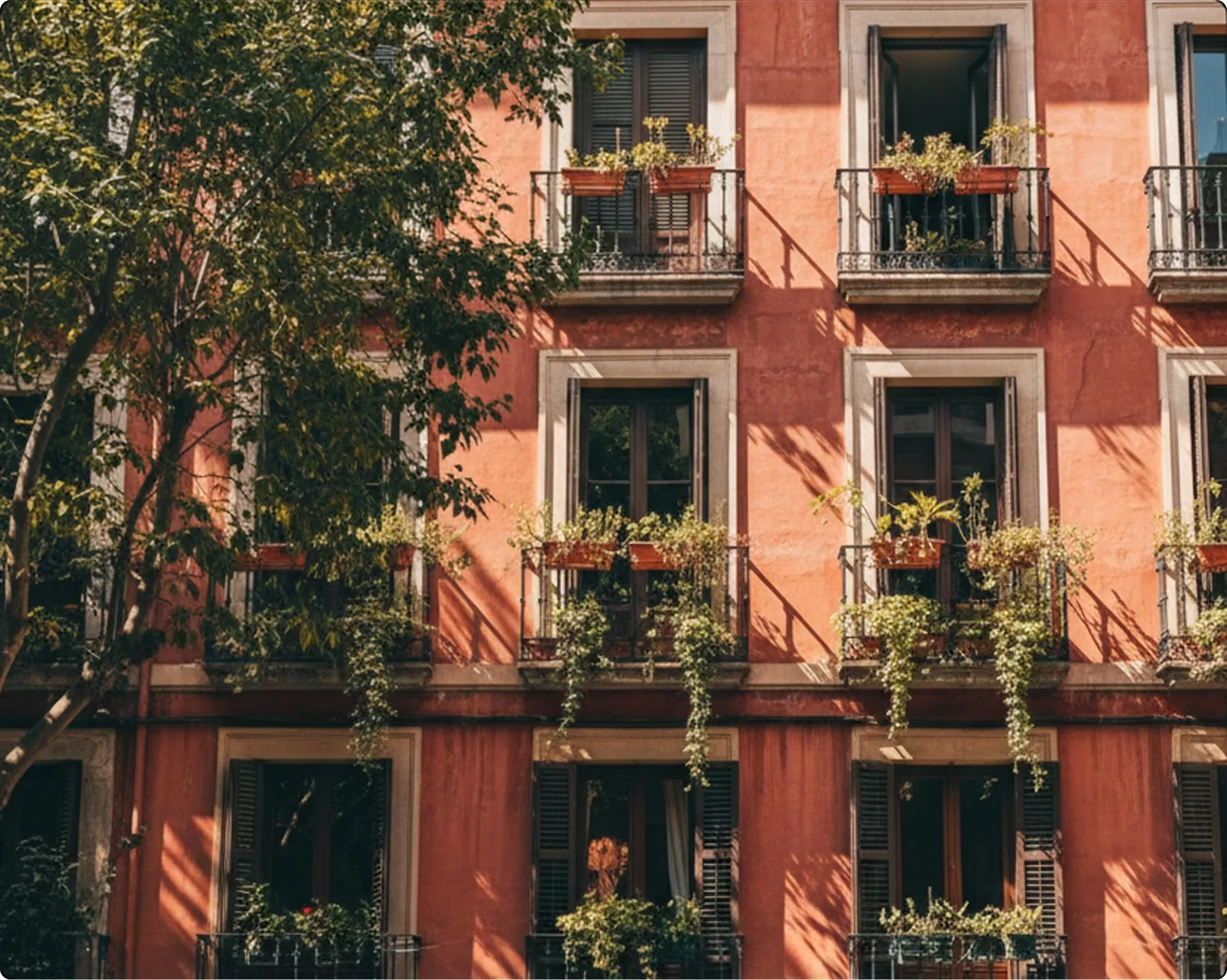Facade of a terracotta-red apartment building with wrought iron balconies overflowing with green plants, sunlit urban architecture