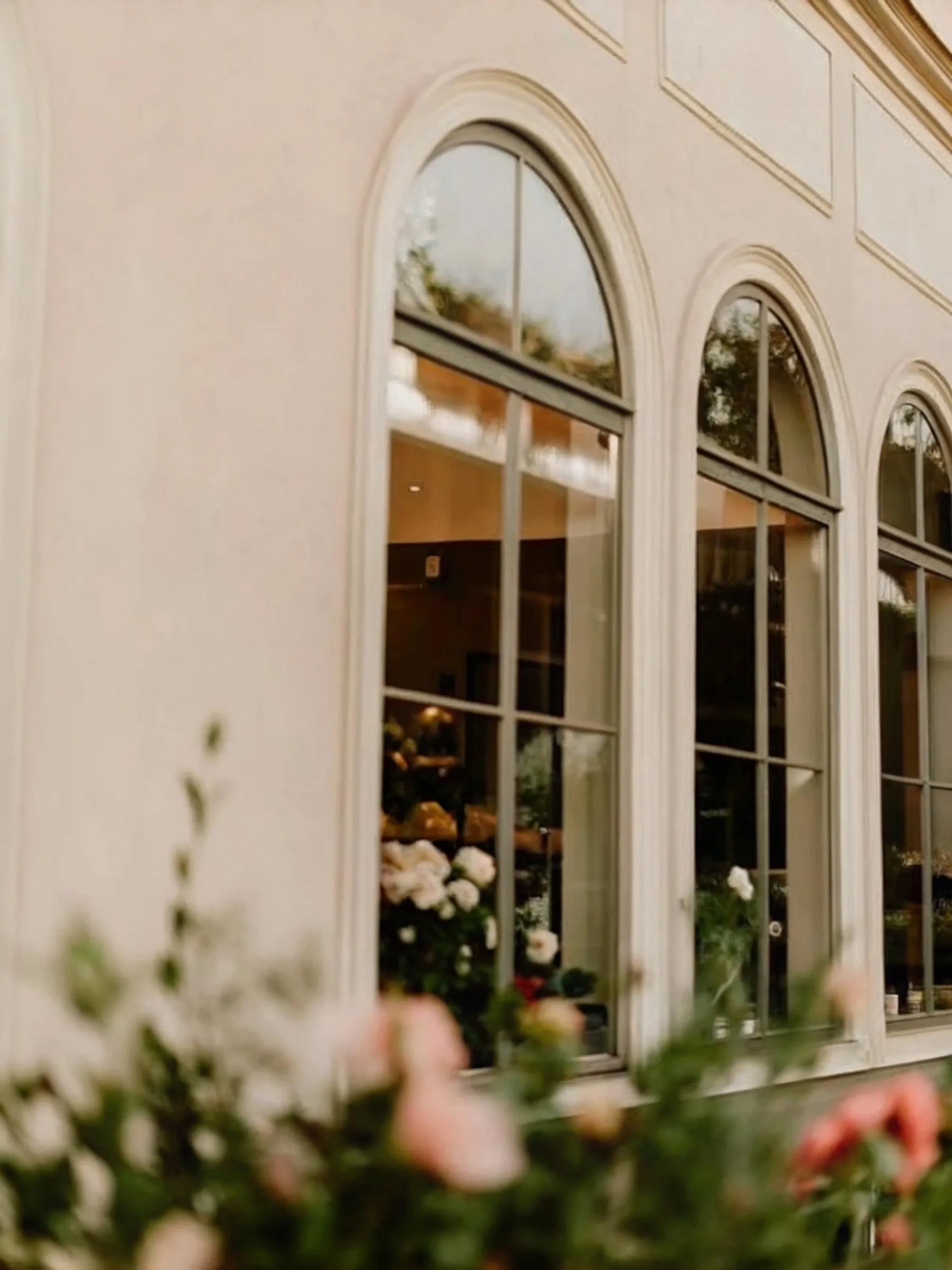 Tall arched windows with dark metal frames on a cream-colored building exterior, with blurred pink roses in the foreground