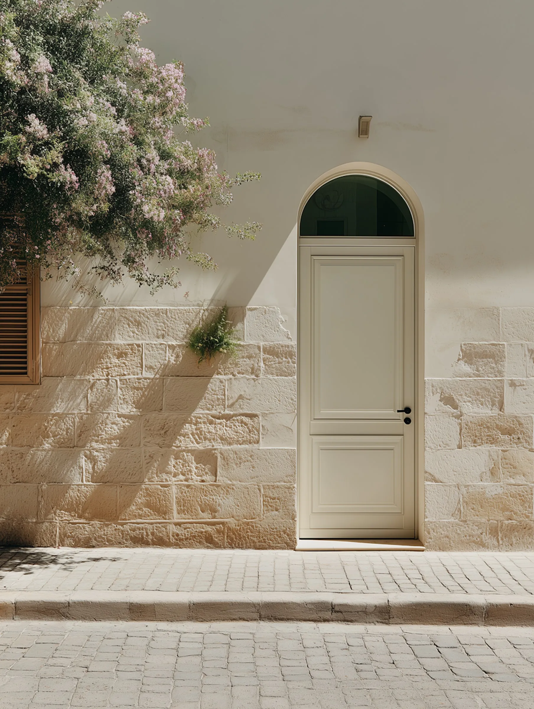 Arched cream-painted door set in a limestone block wall, with a flowering tree casting diagonal shadows on the facade, Mediterranean street architecture